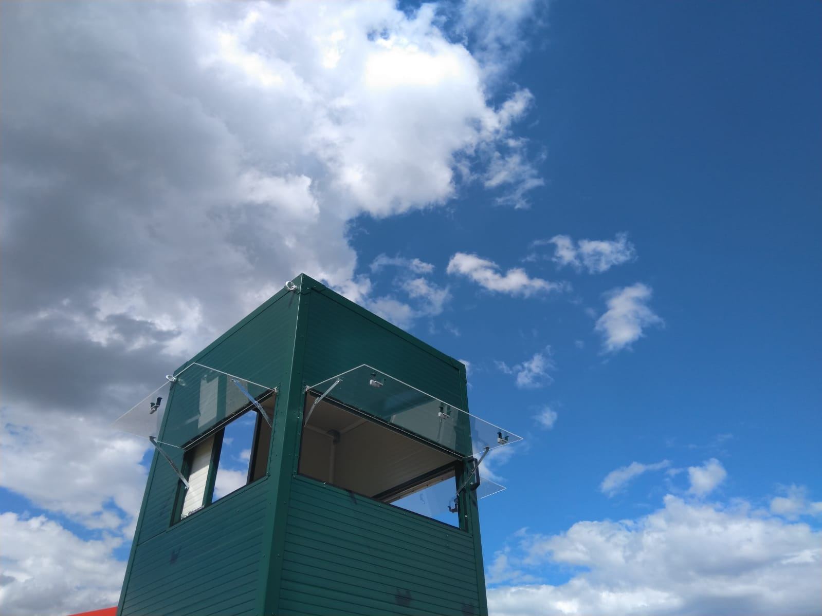 oberer Teil einer grünen mobilen Jagdkanzel mit 2 nach oben geöffneten Fenstern. Vor blauem Himmel mit Wolken