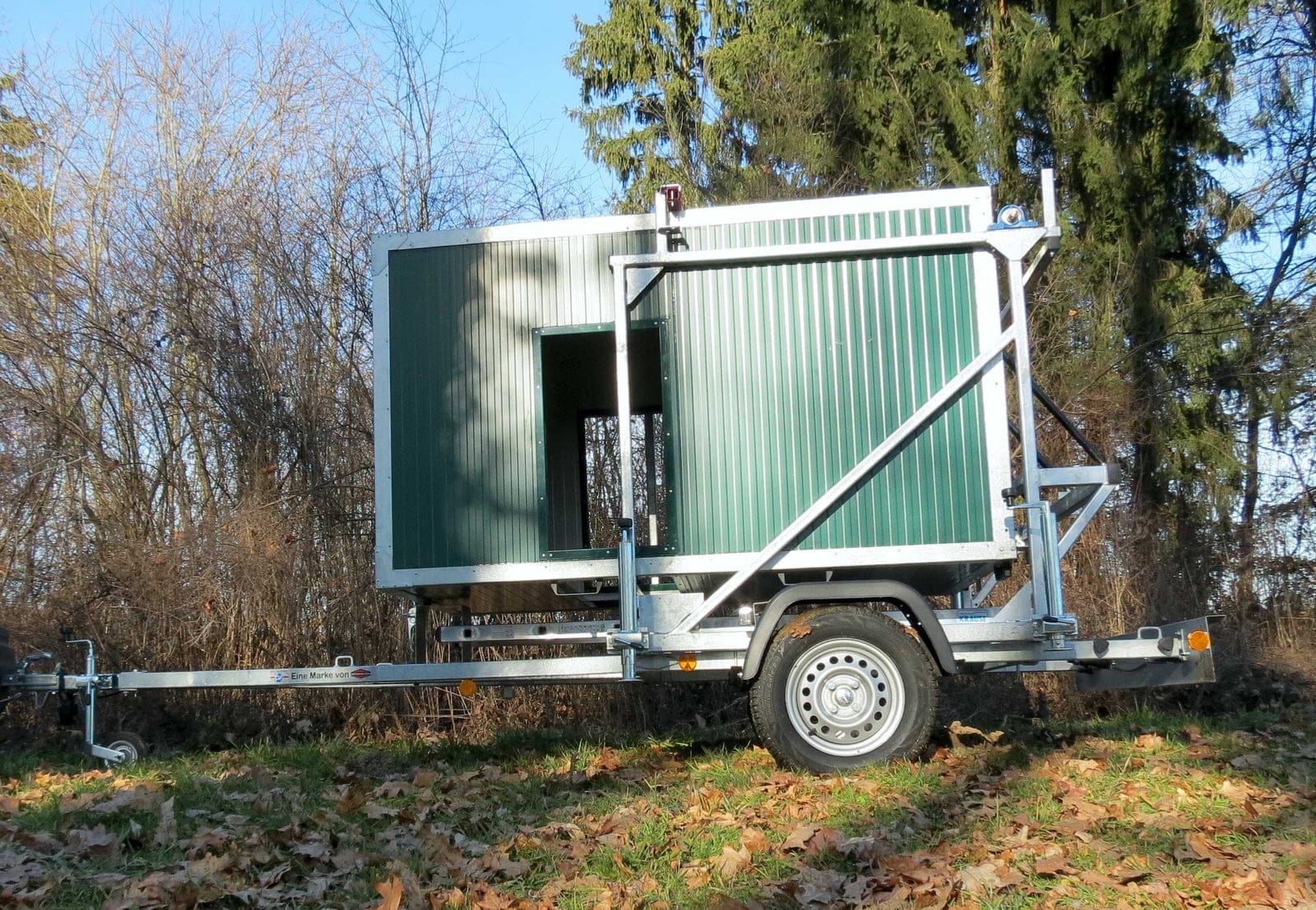 grüne mobile Jagdkanzel, zusammengeklappt auf einem Trailer, in der Mitte des Bildes auf Rasen, mit viel Laub im Vordergrund und kahlen Bäumen im Hintergund